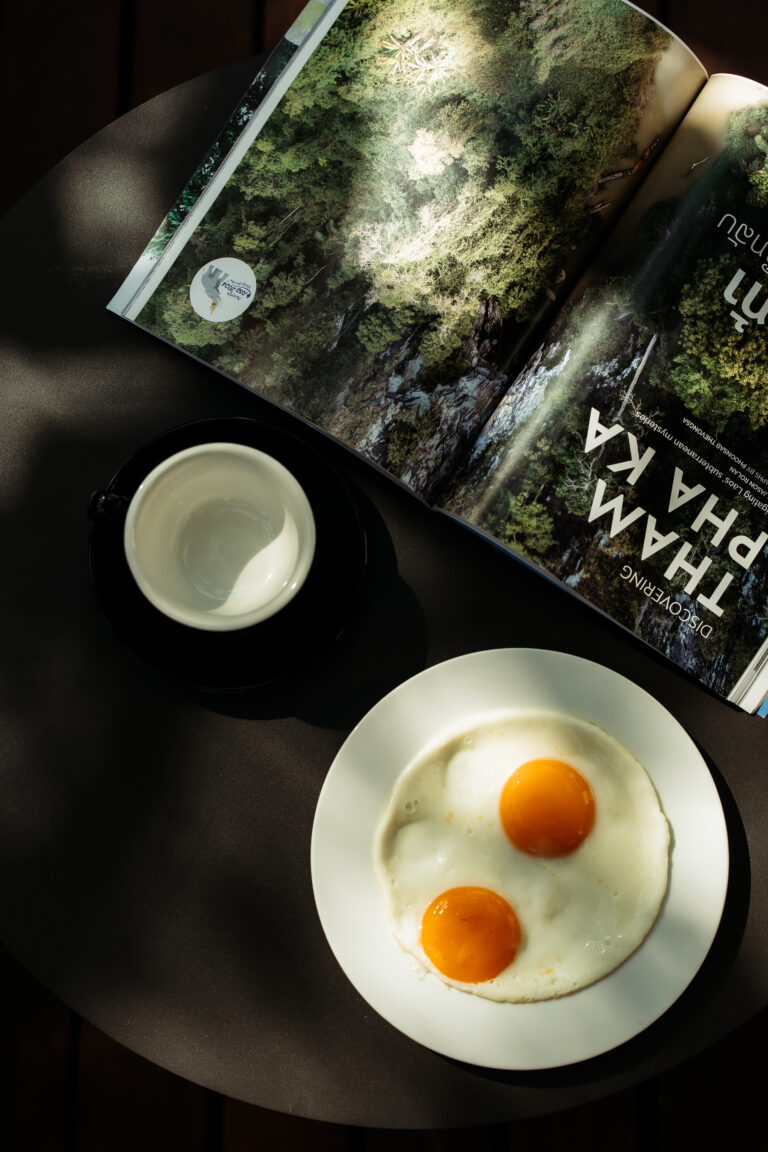 A peaceful breakfast scene at Athena Hotel Pakse, featuring fried eggs and coffee alongside a magazine about local travel.