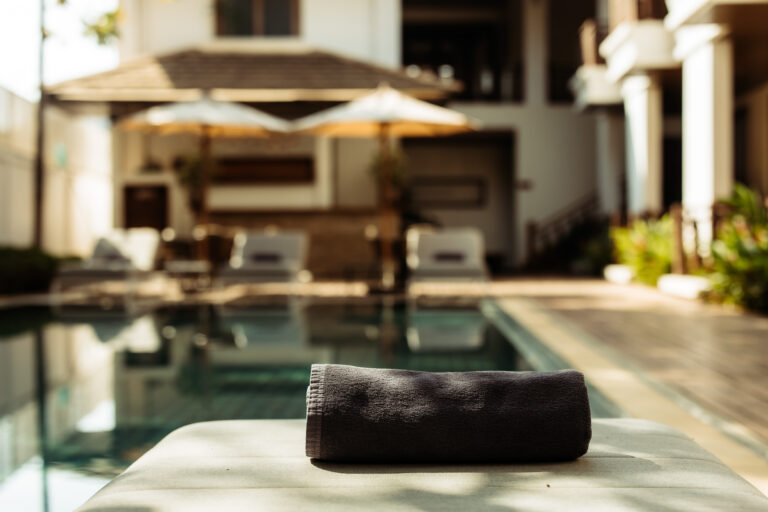 A close-up of a fresh towel ready for a guest, with the beautiful swimming pool and lounge area of Athena Hotel Pakse softly blurred in the background.