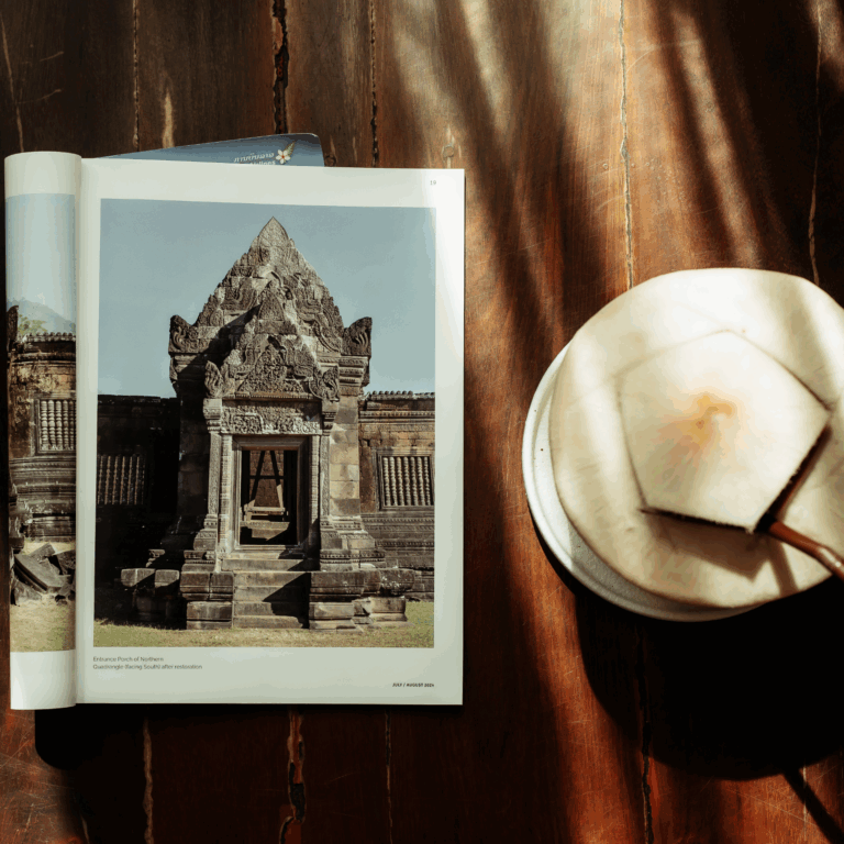 A refreshing coconut drink served on a wooden table next to an open magazine showing a photo of ancient temple ruins in Laos.